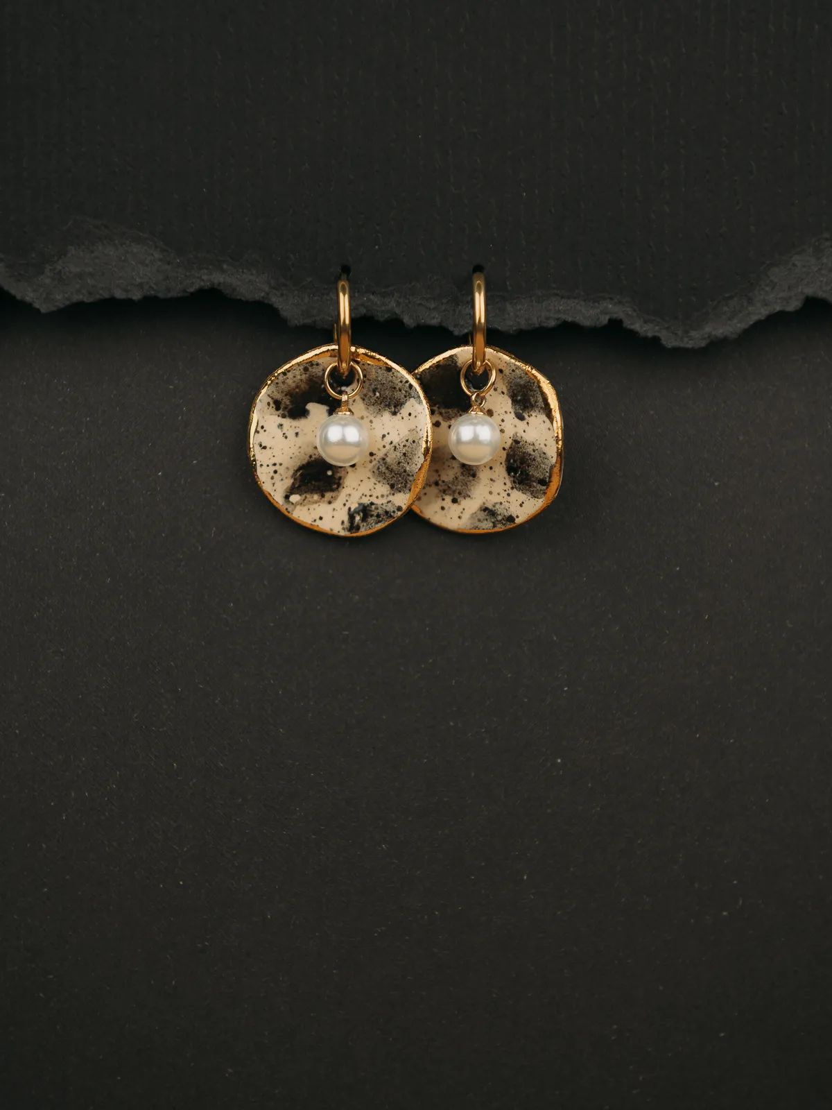 Two gold-hoop earrings with speckled cream and black enamel discs and small pearl drops hang against a dark textured background with a torn paper edge above. The product shot is styled with soft, moody lighting that highlights the jewelry in a commercial product photography aesthetic.