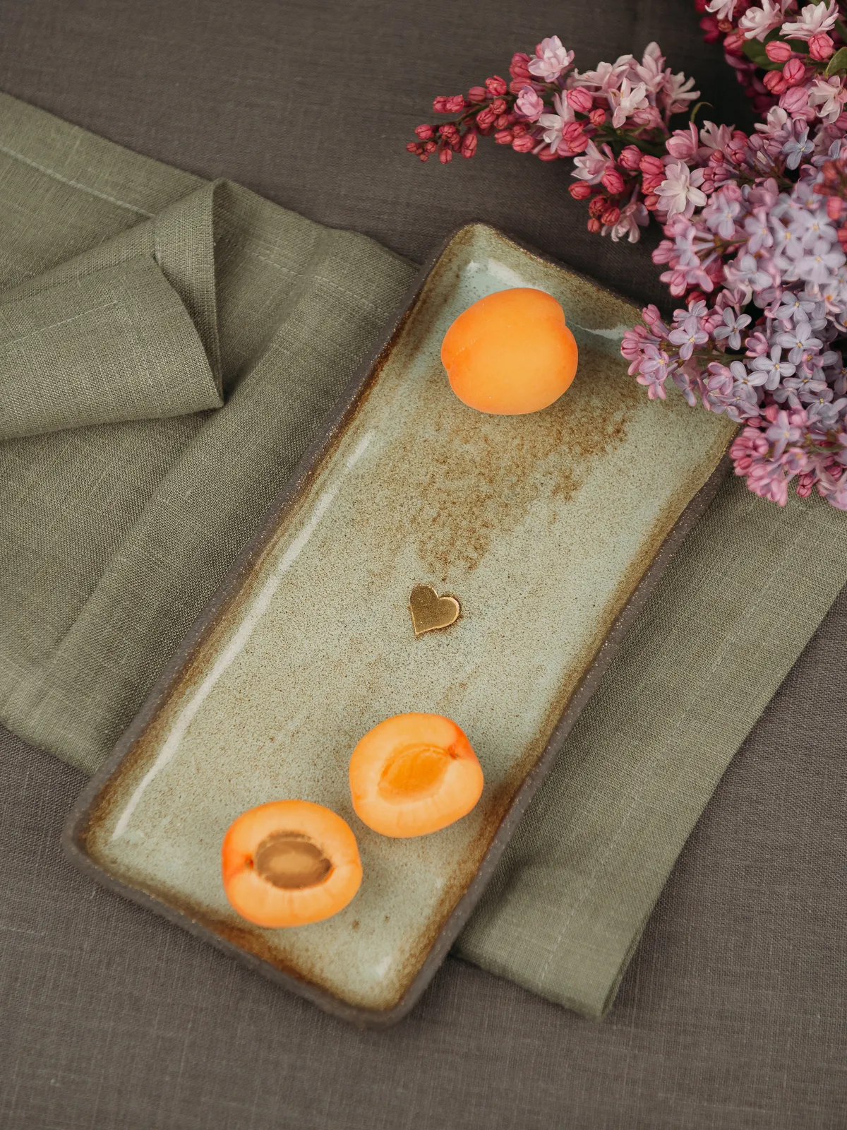 A still life product-style shot shows a rectangular rustic ceramic tray with a small heart detail on an olive linen napkin atop a brown fabric surface. An apricot sits whole at the top and two halved apricots at the bottom of the tray, with purple-pink lilac blossoms in the upper right;