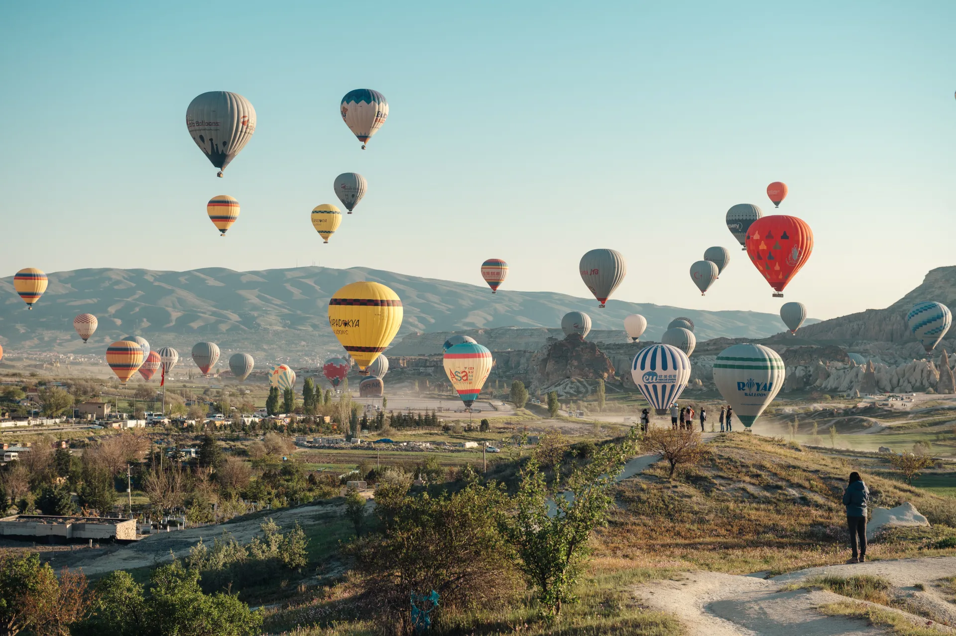 Stambula/Kapadokija — Numerous colorful hot air balloons float above a rocky, hilly landscape at sunrise, with a few people standing on a grassy ridge in the foreground watching the scene.