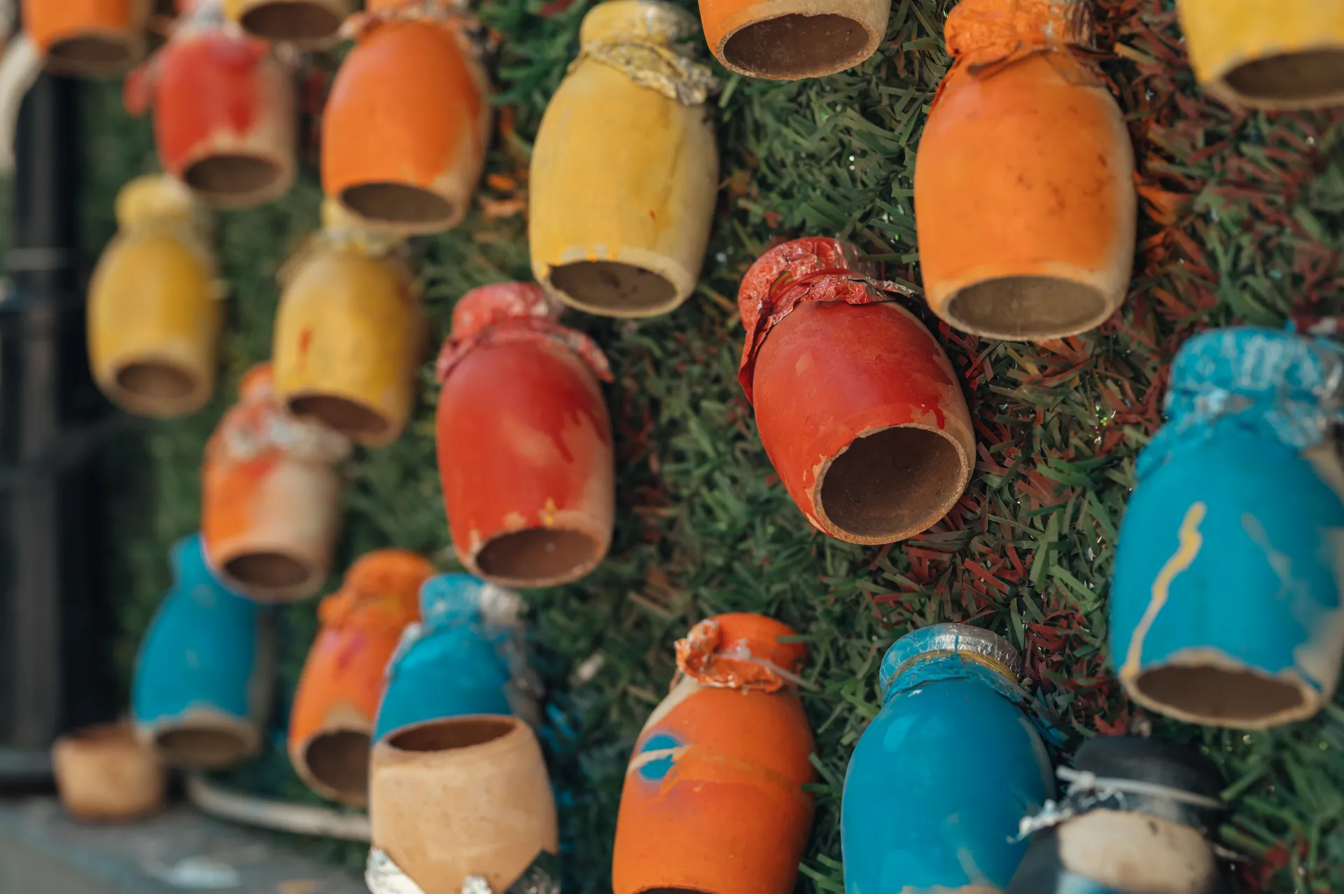 Stambula/Kapadokija — A close-up shows small hand-painted clay pots in red, orange, yellow and blue mounted upside-down on a green artificial grass wall. The ceramic vessels have drips and chipped paint, forming a vibrant still-life street-art installation in soft natural light.