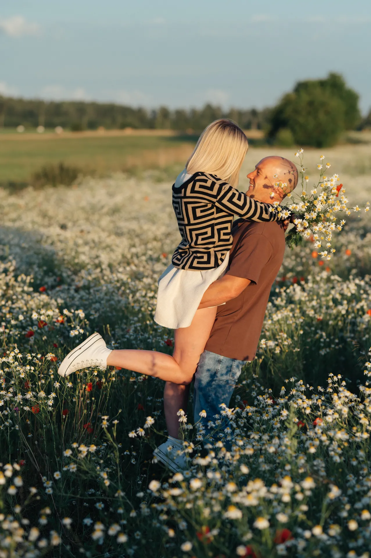 A couple in a sunlit field of white wildflowers, a bald man in a brown t-shirt and jeans lifts a blonde woman in a black-and-white geometric sweater and white skirt; she holds a bouquet of daisies. The golden late-day light and romantic outdoor pose suggest a lifestyle portrait or couple’s photoshoot.