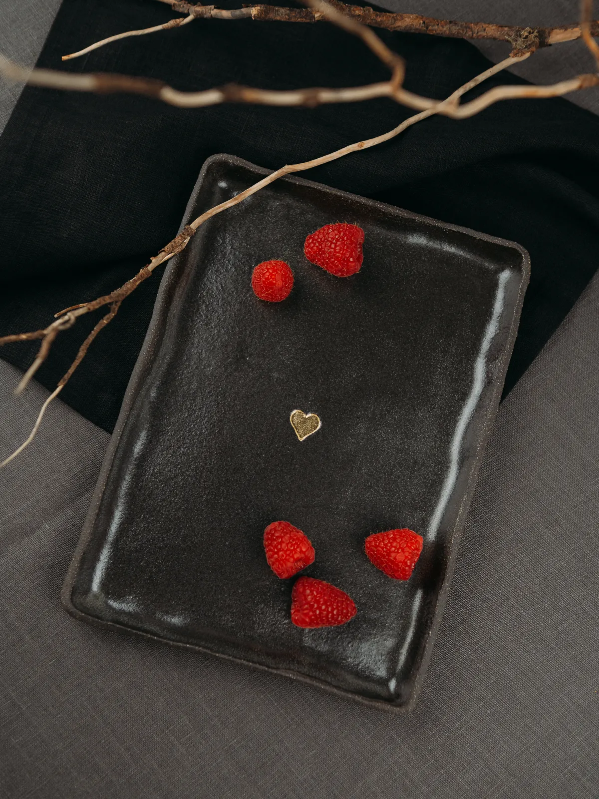 A dark rectangular ceramic plate holds five red raspberries and a small gold heart, set on gray fabric beneath bare branches in a moody still life composition.