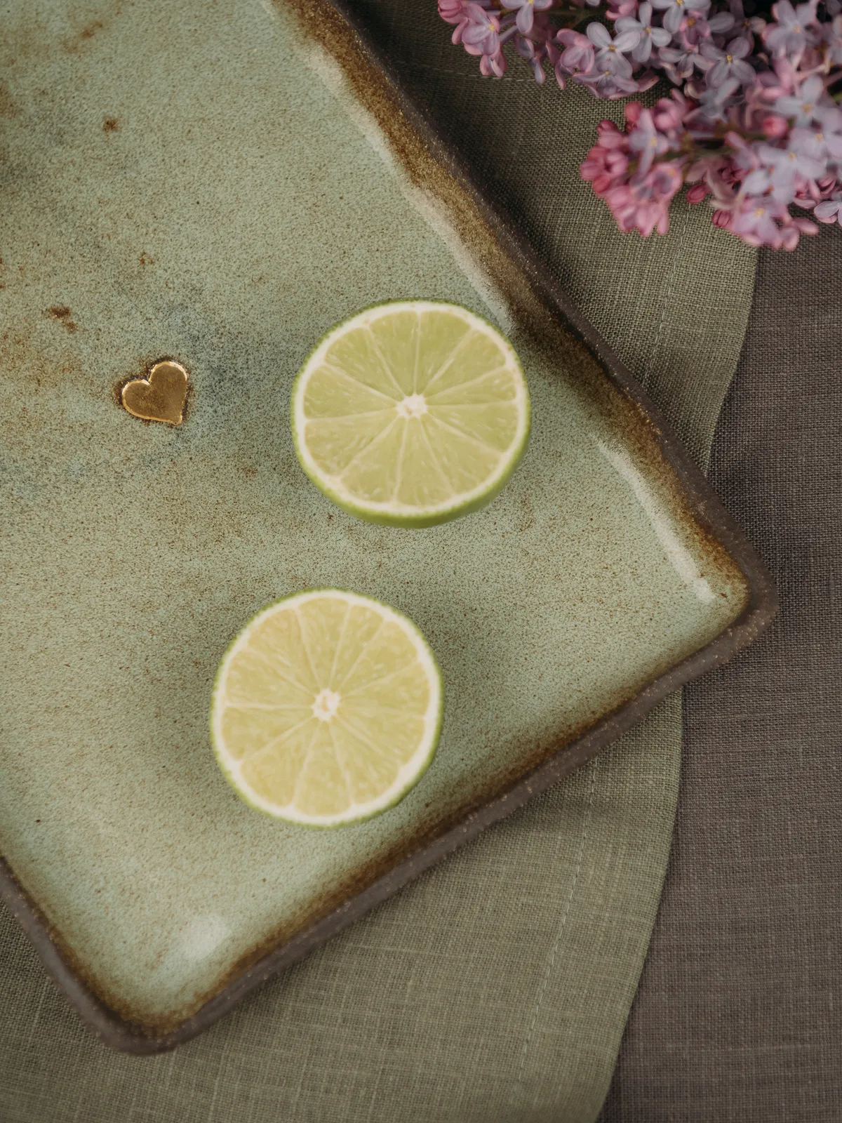 A top-down still life shows two lime slices on a rectangular sage-green ceramic plate with a small gold heart accent, placed on a linen cloth with lilac blossoms in the upper right. Soft natural light creates a calm, minimal product-style composition.