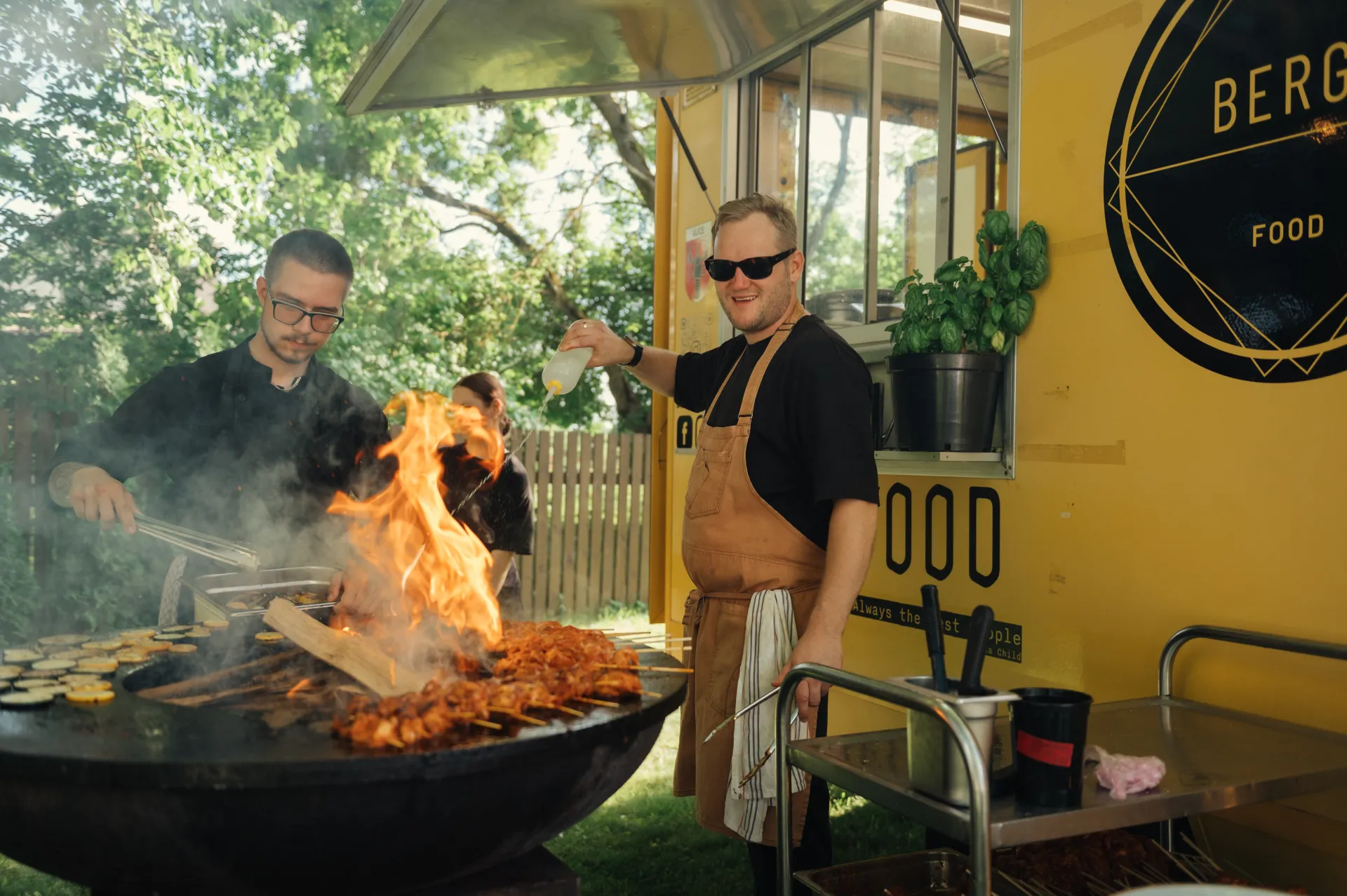 Two chefs in black shirts work over a large outdoor grill with open flames and skewered meat beside a bright yellow food truck with a potted plant on the service window. One chef uses tongs on the fire while the other smiles and sprays seasoning, in daylight with trees and a wooden fence behind them.