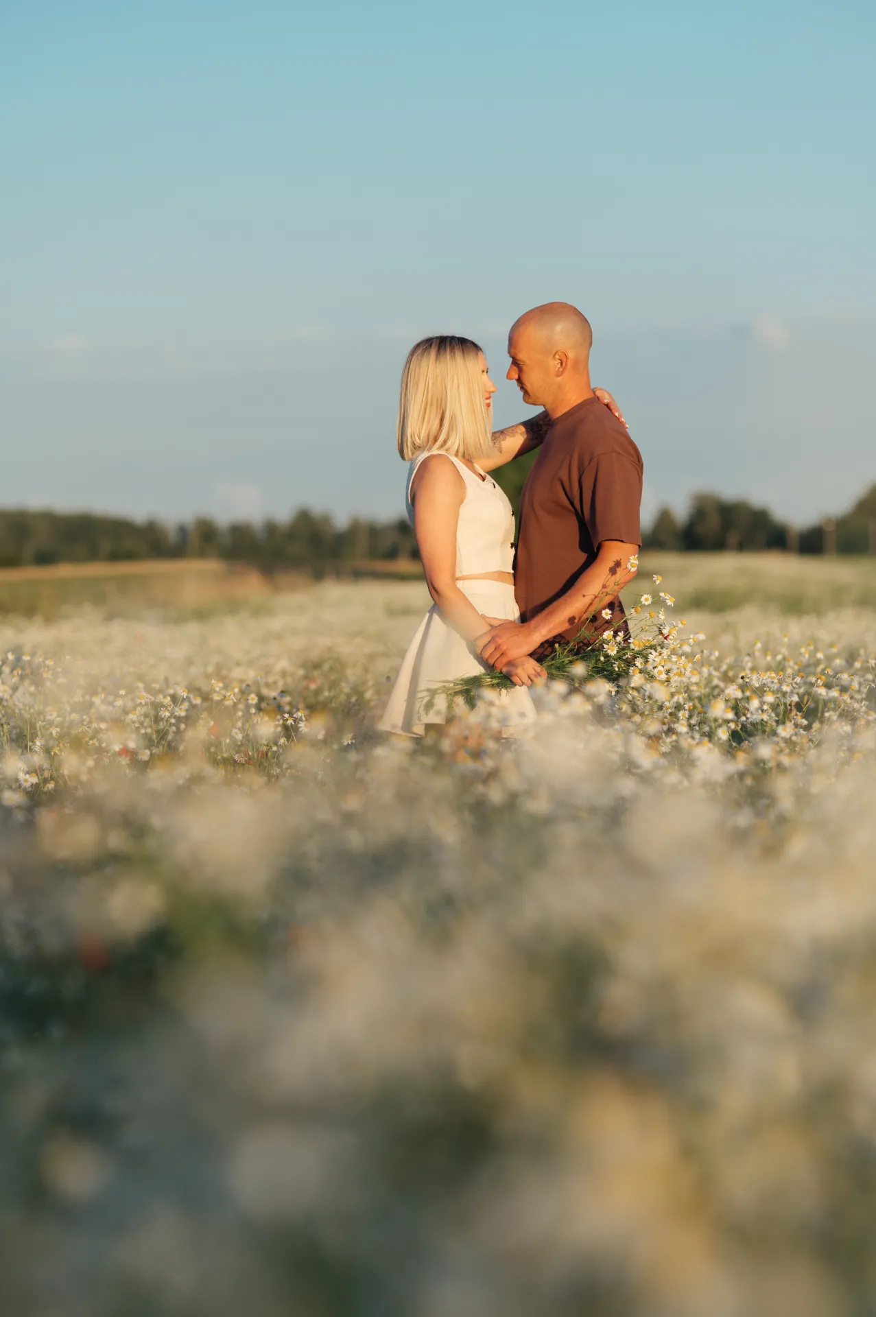 A couple stands close in a field of white wildflowers, gazing at each other under a clear sky, captured in a soft-focus portrait style.