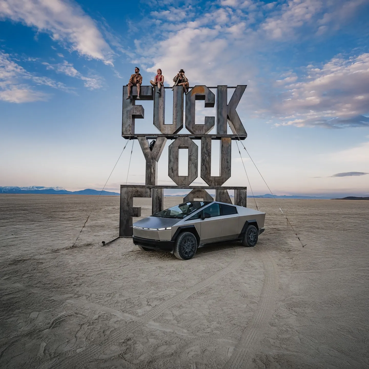 Fuck you Elon — Three people sit atop a large metal sculpture spelling 'FUCK YOU' in a flat desert under a cloudy sky, with a silver angular electric vehicle parked in front; commercial automotive photography.
